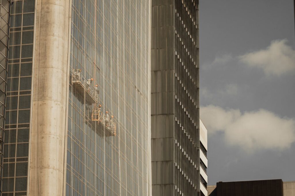 Window cleaners on suspended platforms working on a skyscraper facade against a cloudy sky.